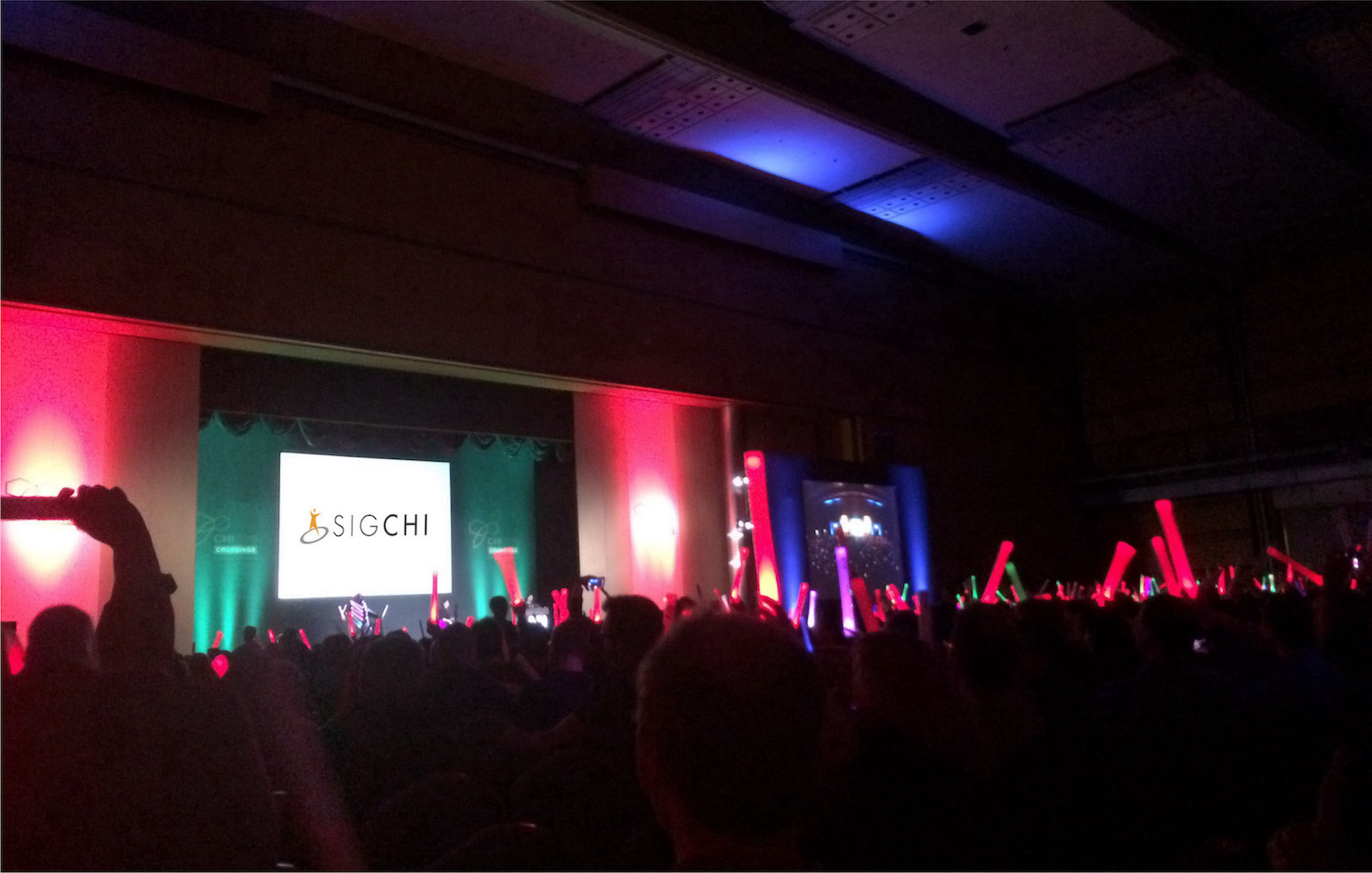 An image of the crowd during a conference keynote. In the distance, there is a stage with a screen that has "SIGCHI" projected onto it.